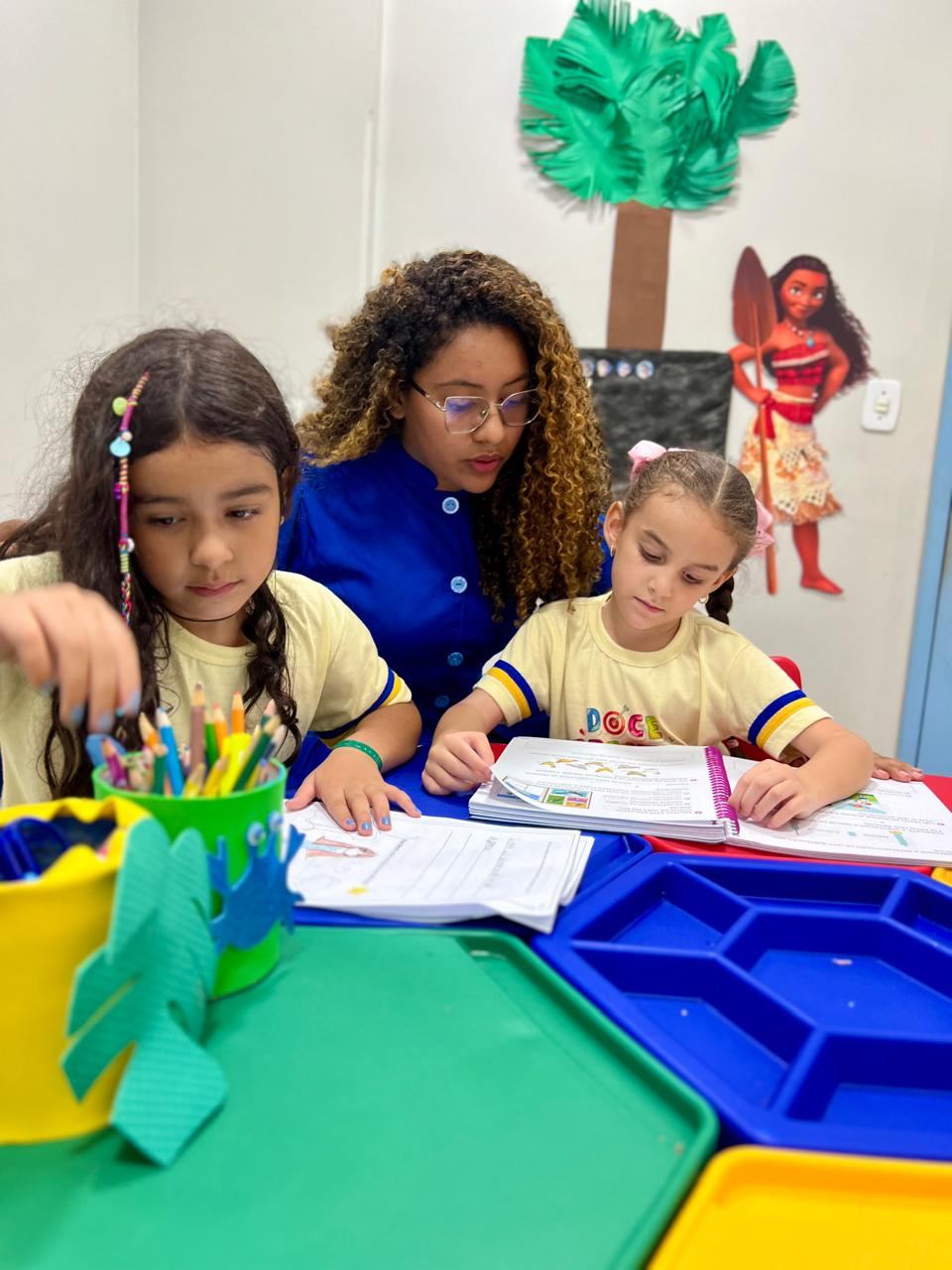 Educadora do Doce Aprender acompanhando duas crianças na lição, com cuidado e atenção individualizada