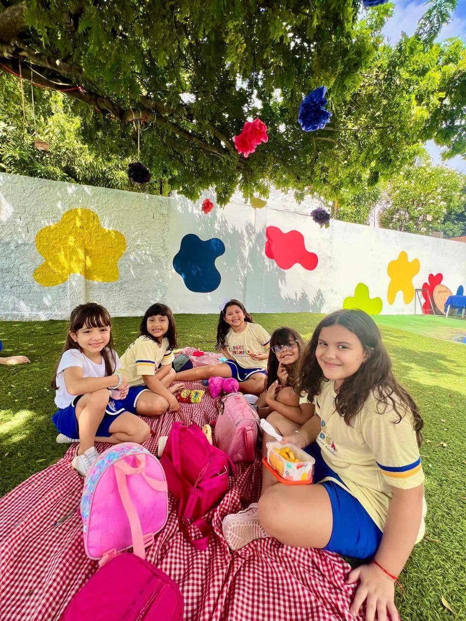 Crianças do Doce Aprender em piquenique no quintal sob a árvore com mural colorido ao fundo